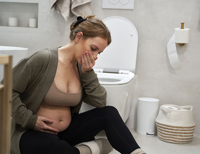 Pregnant woman feeling sick in bathroom, sitting on floor near toilet, holding hand to mouth.