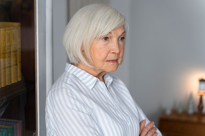 Elderly woman with white hair and striped shirt standing thoughtfully, suggesting a mother-in-law scenario.