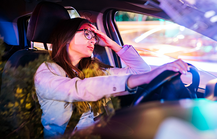 Woman driver looking stressed, resting head on hand, city lights reflecting on car window.