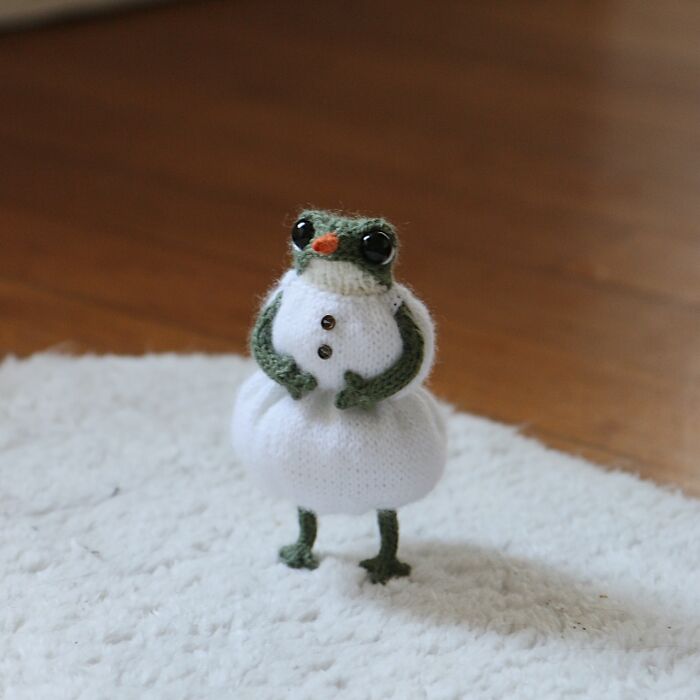 Knitted miniature frog in a white dress, standing on a soft surface.