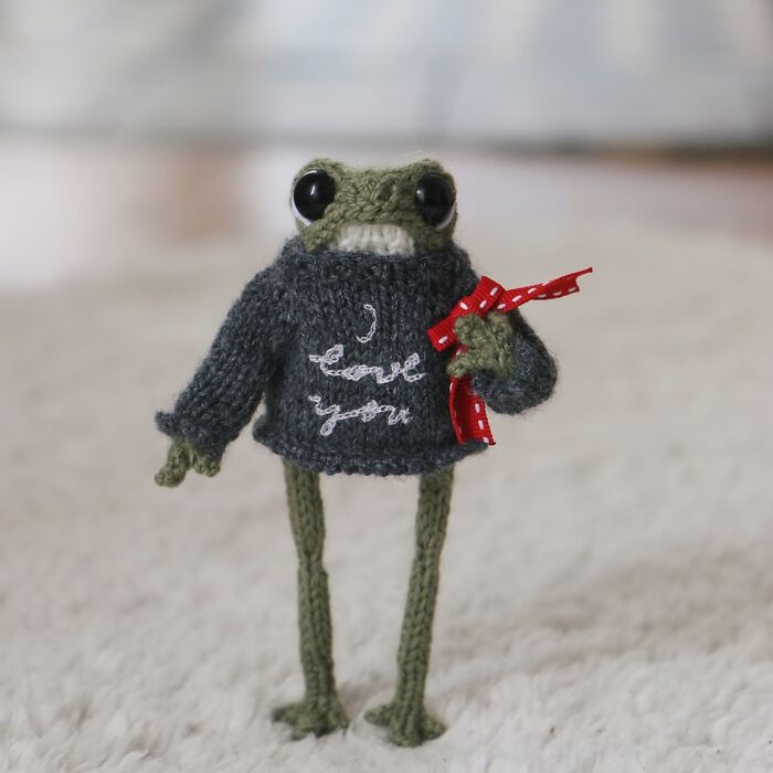 Miniature knitted frog in sweater with "I love you" design, holding a red ribbon, on a soft white background.