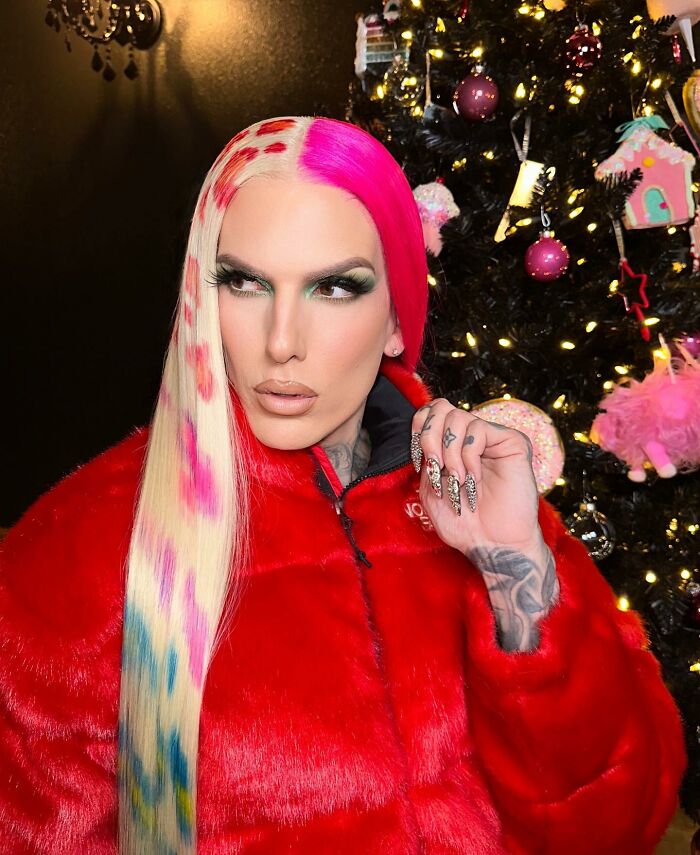 Celebrity in colorful hair and red fur jacket poses by a decorated Christmas tree, festive atmosphere.
