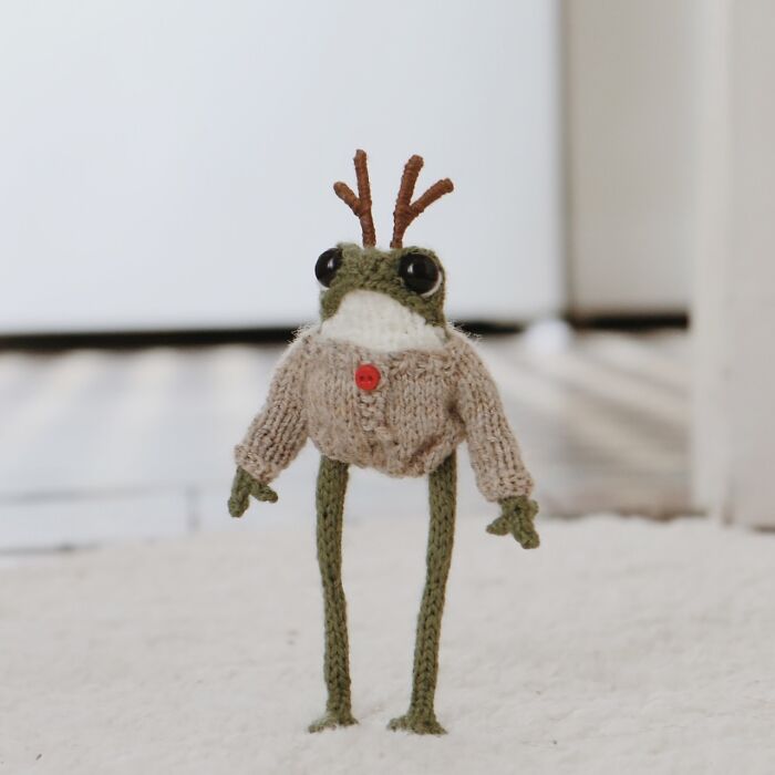 Knitted miniature frog with antler-like adornments, wearing a sweater with a red button, crafted by a French artist.