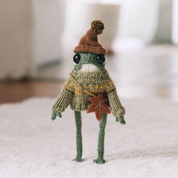 Miniature frog in a knitted sweater and hat, crafted by a French knitter, standing on a white surface.