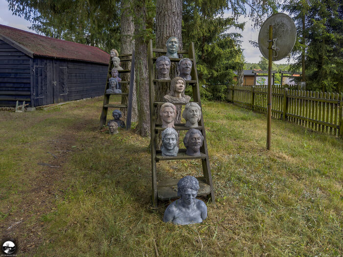 Sculptures by Adam Szubski displayed on wooden ladders in a grassy outdoor setting near a rustic shed and satellite dish.