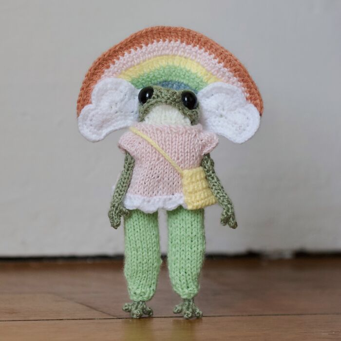 Miniature knitted frog by a French artist, wearing a pastel rainbow and pink outfit, standing on a wooden surface.