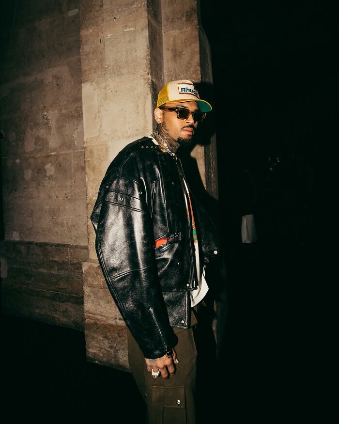 Celebrity wearing a leather jacket and hat in a dimly lit setting.