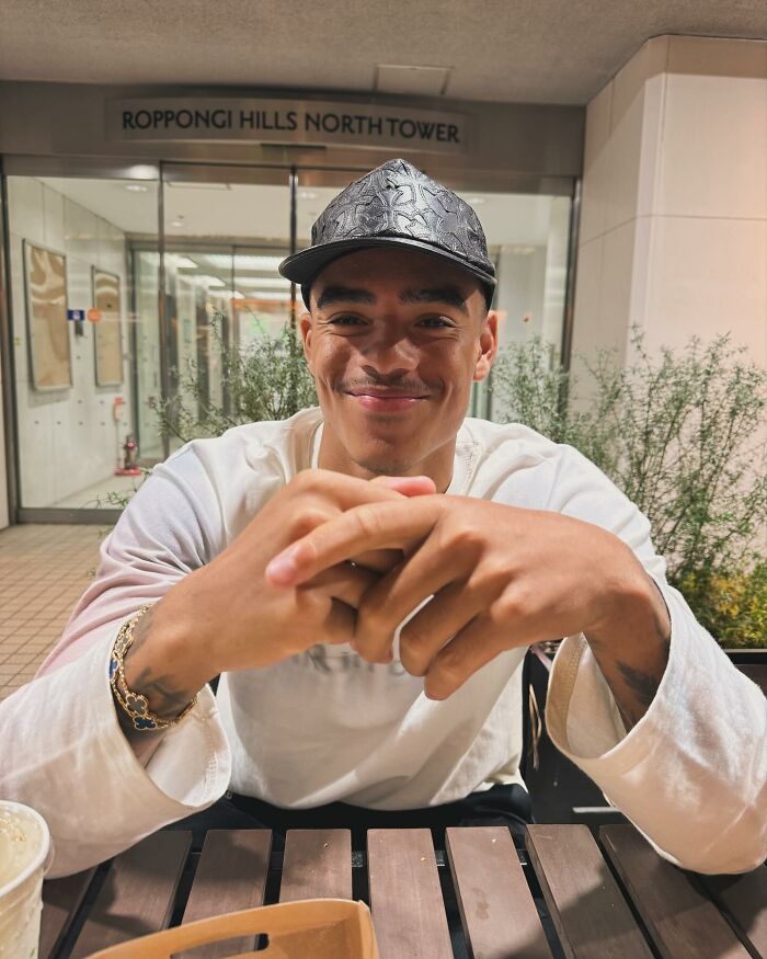 Man sitting at a table, wearing a black cap and white shirt, in front of Roppongi Hills North Tower entrance.