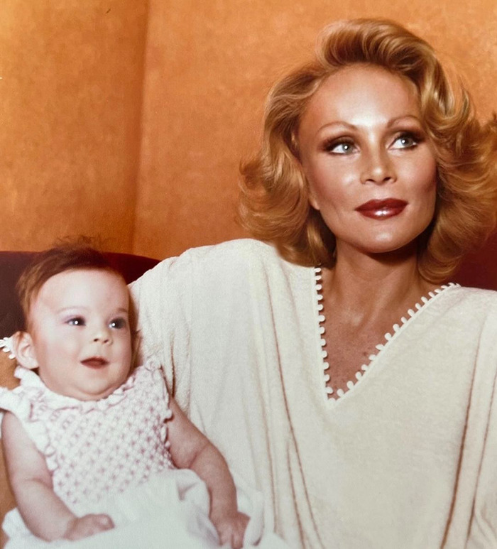 Jocelyn Wildenstein with short blonde hair holds a baby, both smiling, in a warm-colored room. Jocelyn Wildenstein with short blonde hair holds a baby, both smiling, in a warm-colored room.
