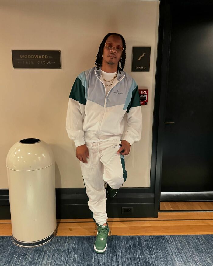 A person in a white tracksuit and green sneakers leans against a wall indoors.