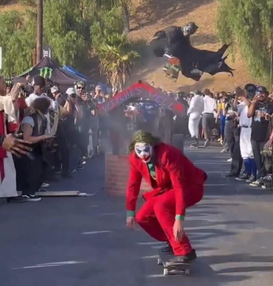 Person in a red suit skateboarding amid a cheering crowd, with another person in a costume jumping in the background.