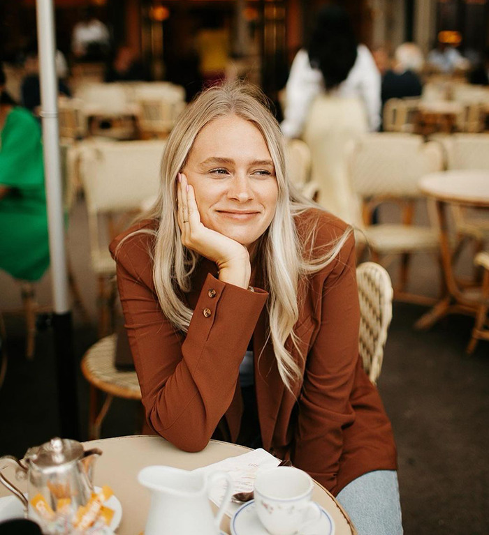 Influencer in a brown jacket smiles at an outdoor caf&eacute; table with a teapot and cup.