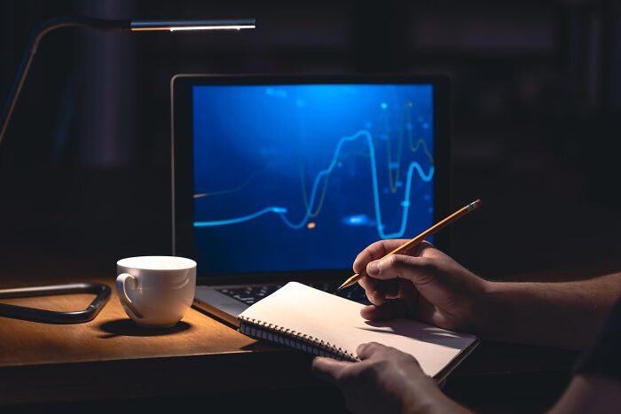 Person taking notes beside a laptop displaying data graphs, symbolizing a risky decision process.
