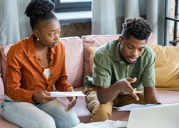 A man and woman discussing financial struggles, sitting on a couch with papers and a laptop.