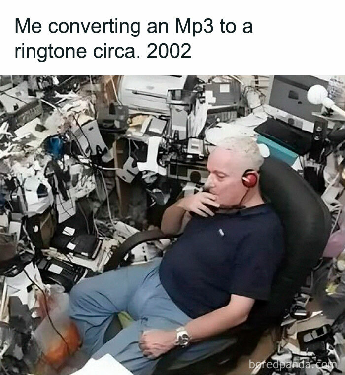 Man in cluttered office using headphones, surrounded by old tech gear, represents IT humor about early 2000s MP3 ringtones.