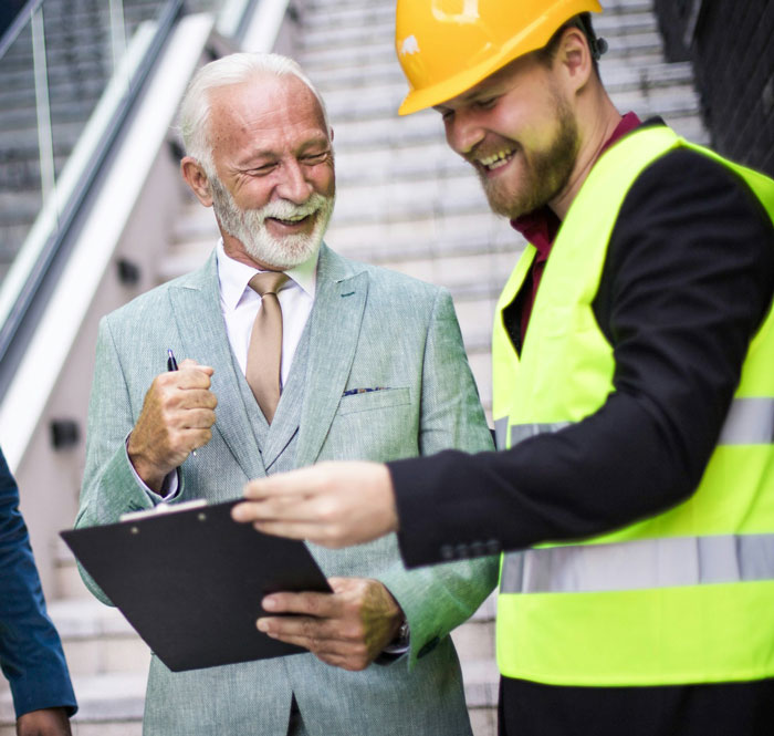 Two men in business attire and safety gear smiling and discussing work outside, highlighting working too fast and malicious compliance. Two men in business attire and safety gear smiling and discussing work outside, highlighting working too fast and malicious compliance.