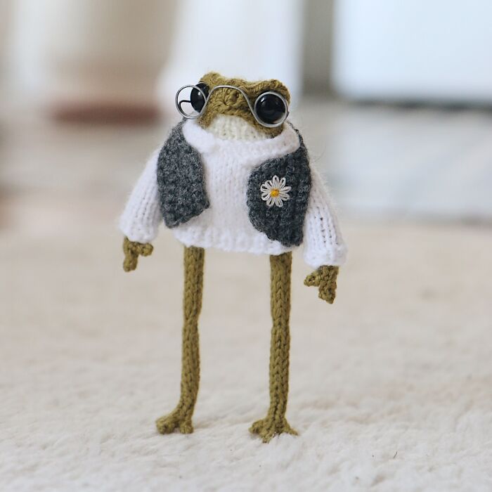 Miniature knitted frog wearing glasses and a white sweater with a flower detail, created by a French knitter.