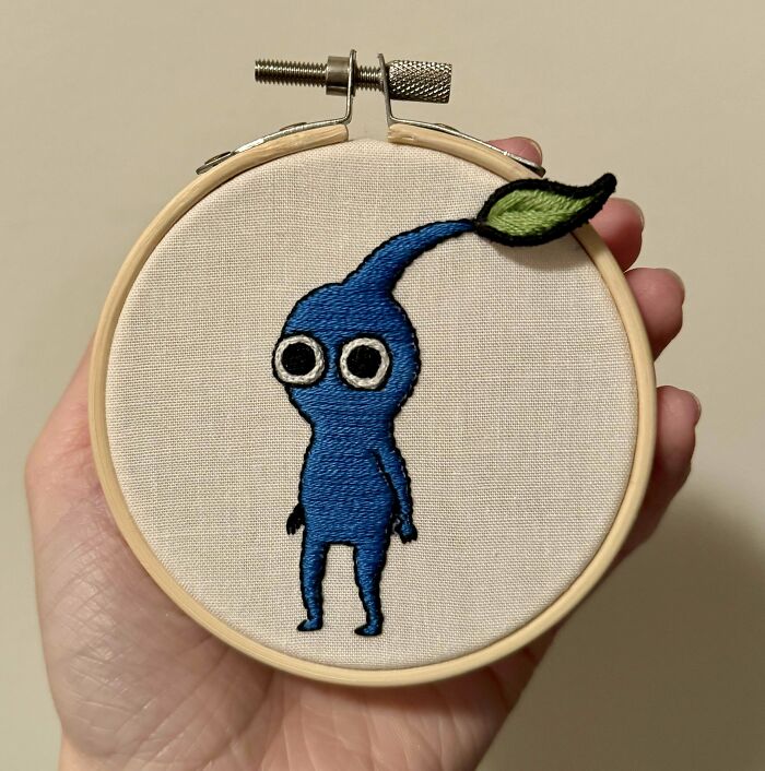 Hand holding an embroidery hoop with a blue character design and a green leaf, showcasing beautiful embroidery designs.