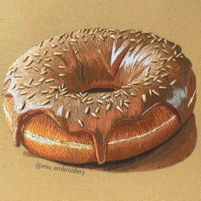 Realistic embroidery design of a chocolate-glazed donut with sprinkles on a tan fabric background.