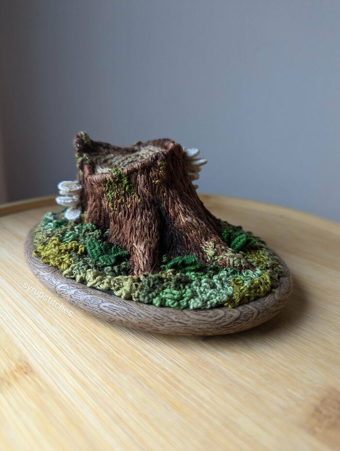 Intricate embroidery design of a tree stump with green foliage on a wooden surface, showcasing beautiful embroidery designs.