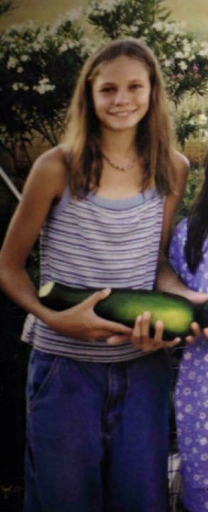 Young person in a striped tank top holds a large zucchini, capturing a blunder years moment outdoors.