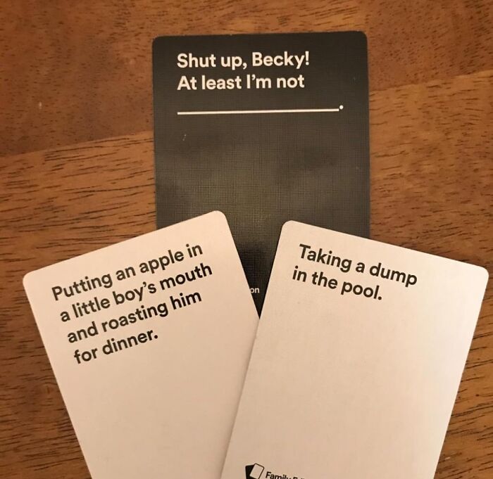 Cards from a humorous adult party game displayed on a wooden table about roasting and pool pranks.