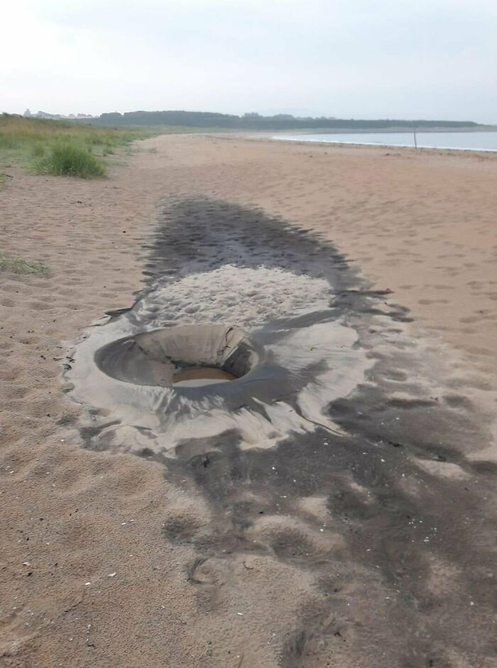 Quirky treasure: A water-filled hole surrounded by dark sand on a beach.