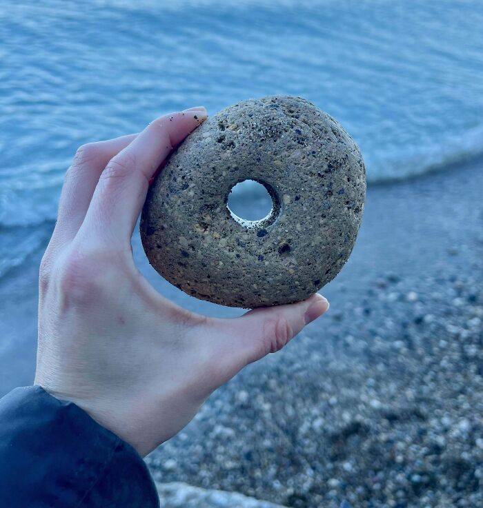 Hand holding a donut-shaped stone, a quirky treasure found washed ashore on a pebbly beach.