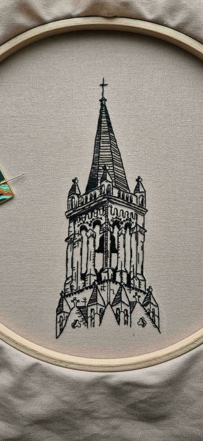 Black thread embroidery design of a detailed church steeple in a wooden hoop on beige fabric.