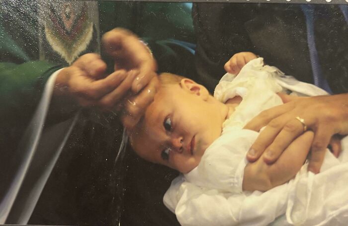 Baby in white gown surrounded by hands, resembling a Renaissance painting moment.