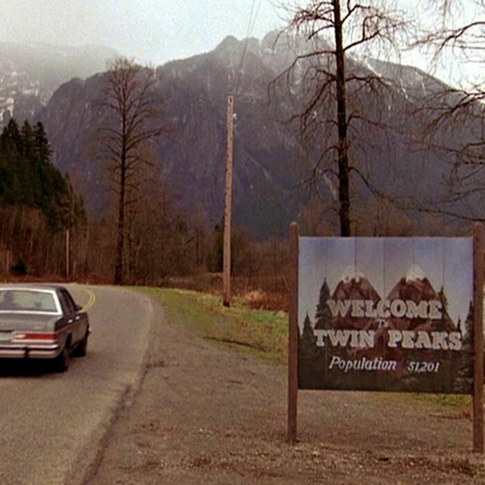 Roadside sign for Twin Peaks with a car approaching in a ‘90s scene.