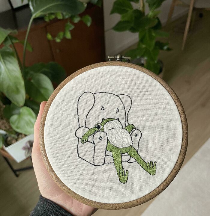 Hand holding embroidery hoop with a stitched frog lounging in an armchair, showcasing beautiful embroidery designs.