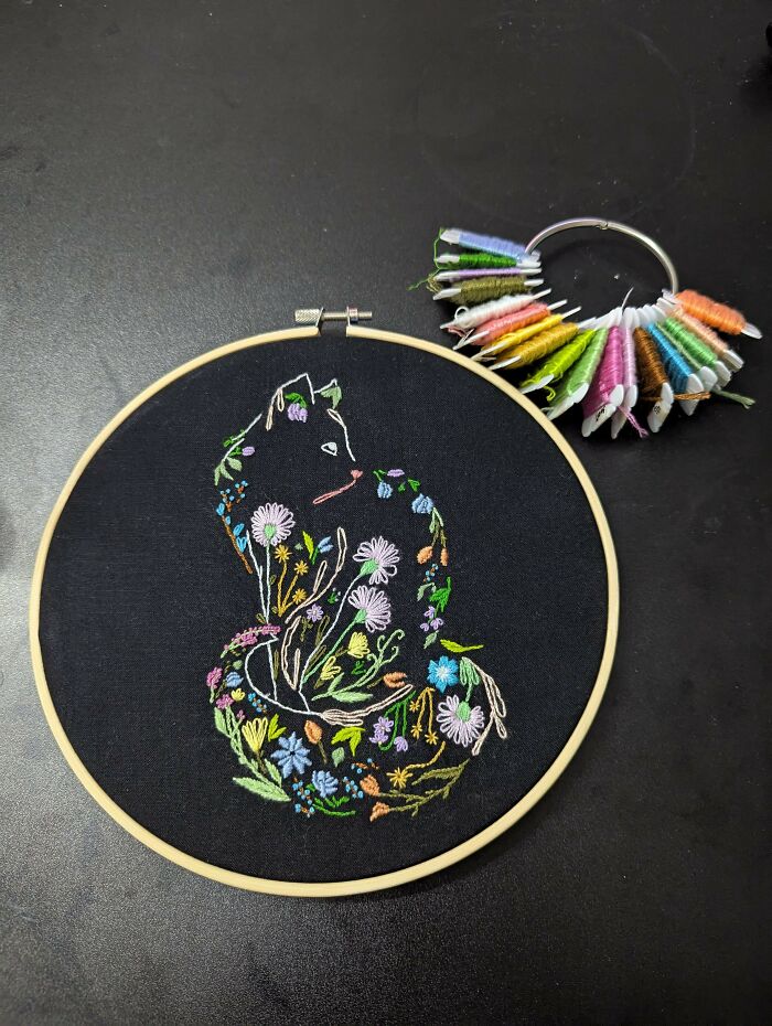 Colorful embroidery designs of flowers and leaves forming a cat shape in a wooden hoop on black fabric.