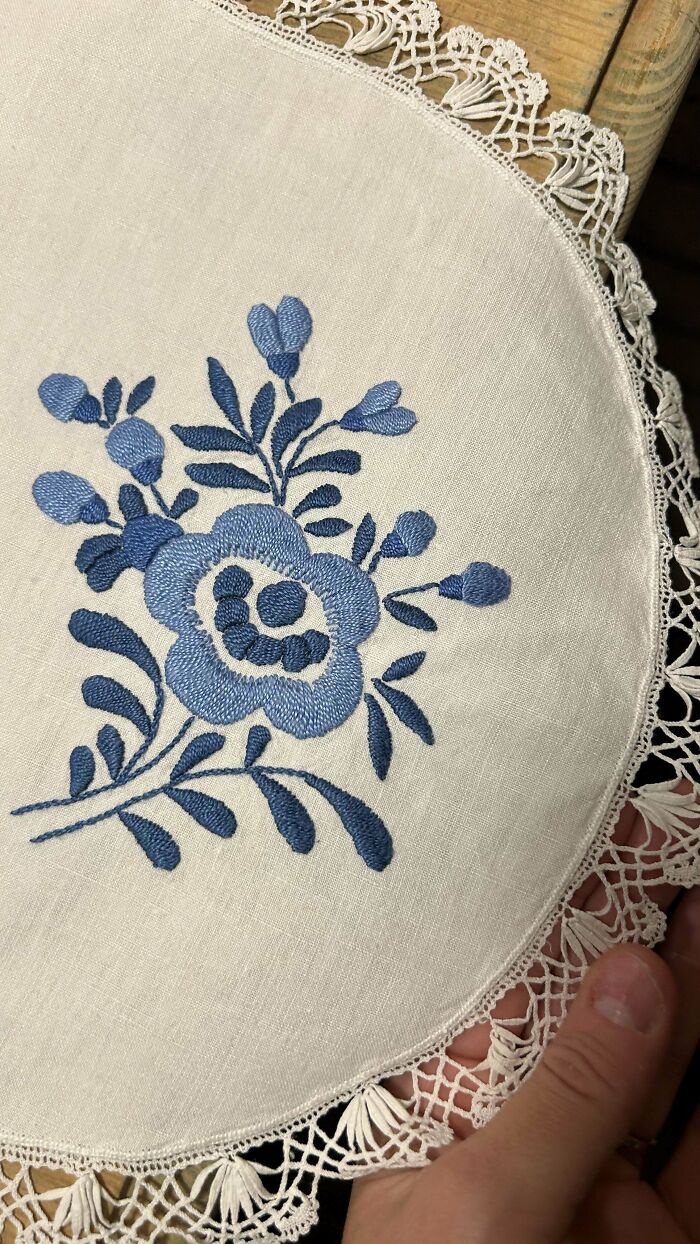 Blue floral embroidery design on white fabric with intricate lace edges held by hand, showcasing beautiful embroidery designs.