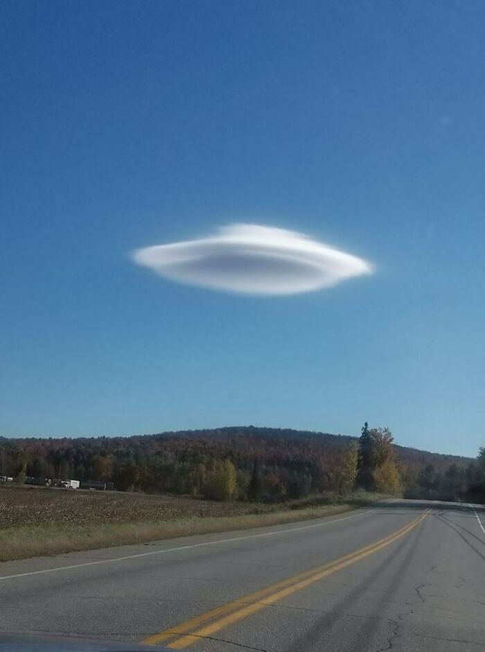 Unusual UFO-shaped cloud hovering in a clear blue sky over a rural road and landscape.