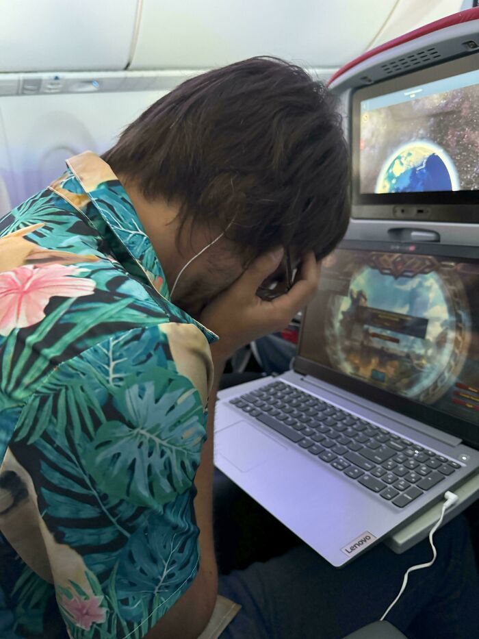 Person regretting a purchase, sitting on a plane with a laptop, wearing a tropical shirt, hands covering face.