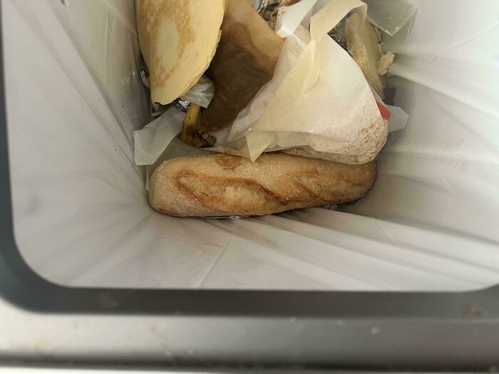 A partially eaten baguette and other discarded food items in a trash bin, highlighting wasteful habits.