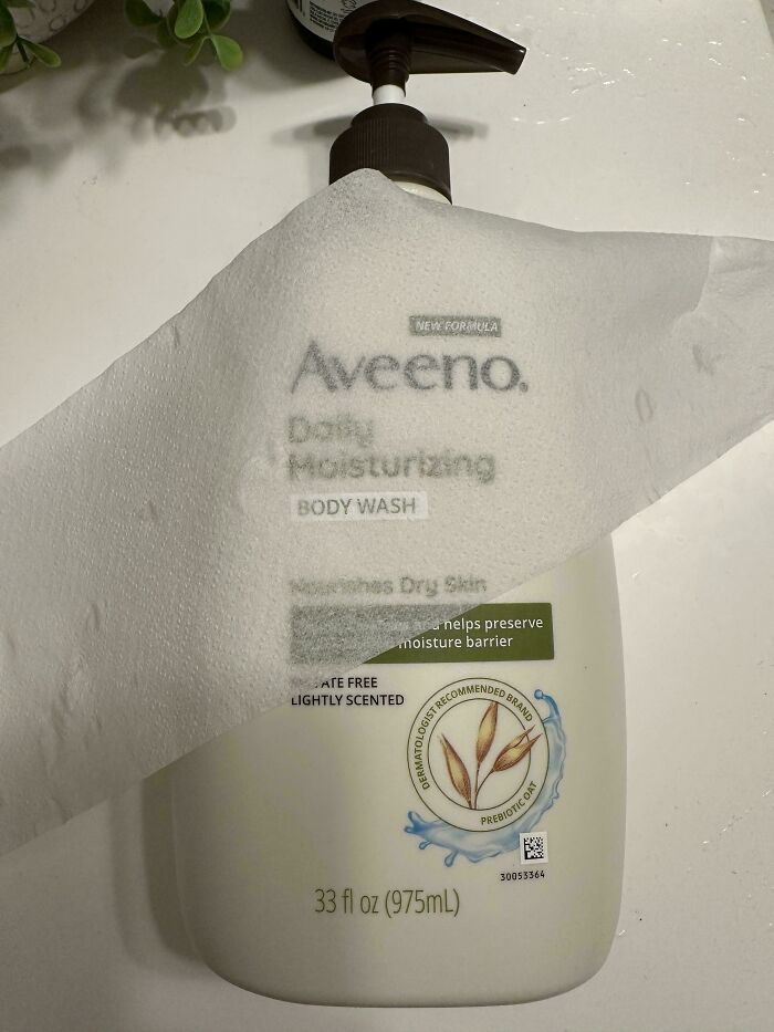 Aveeno body wash with pump wrapped in tissue paper, surrounded by green leaves, possibly a prank by infuriating partners.