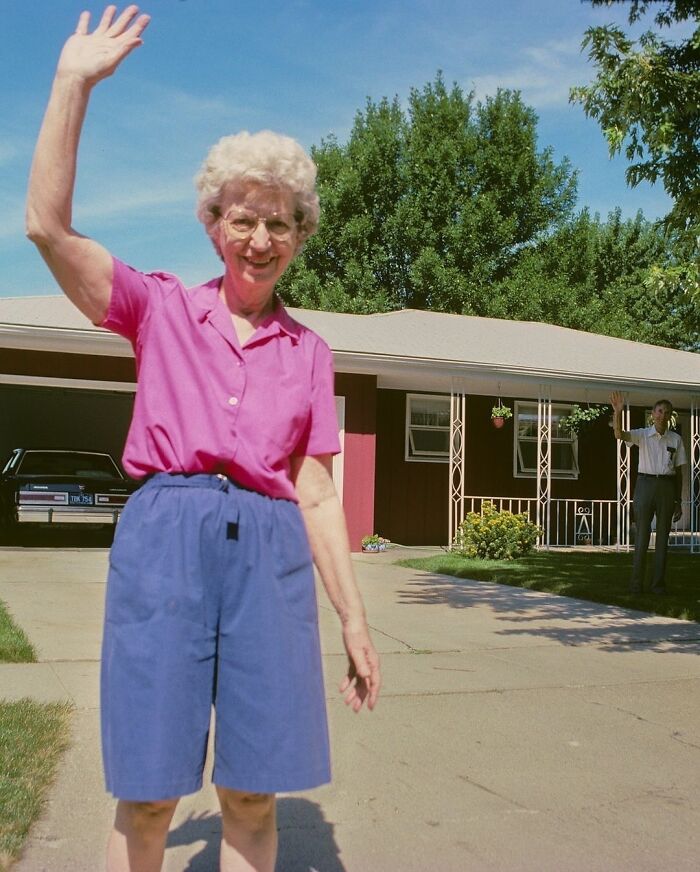Elderly woman waving in '90s driveway scene with a car and man in the background.