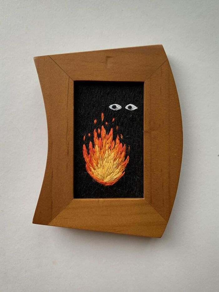 Small framed embroidery design featuring a detailed flame with eyes on a black fabric background.