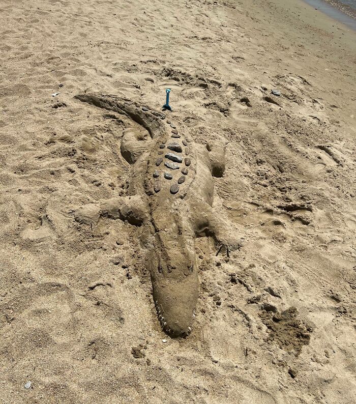 Sand sculpture of an alligator adorned with rocks and a toothbrush on a beach, showcasing quirky treasures washed ashore.