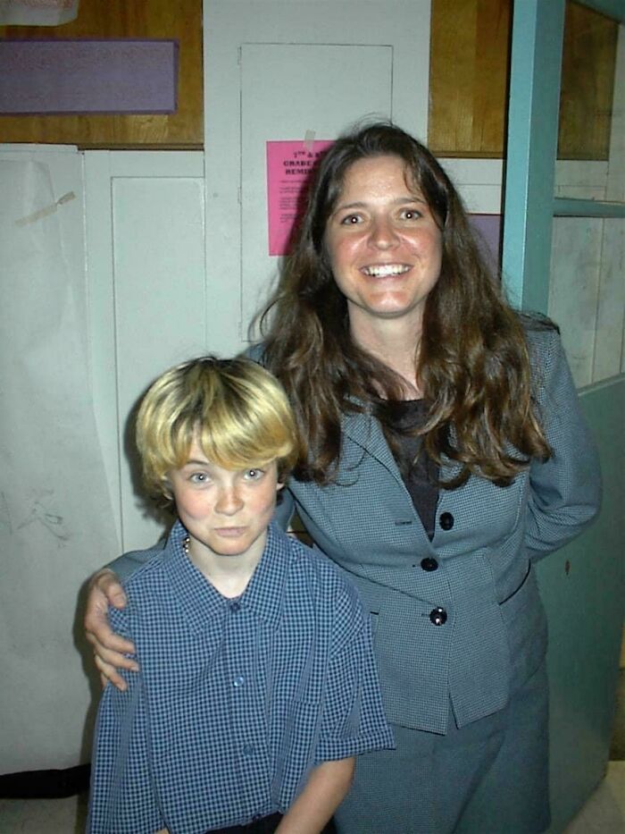 A young boy with blonde hair stands next to a woman in a suit, both smiling, capturing a "blunder years" moment.