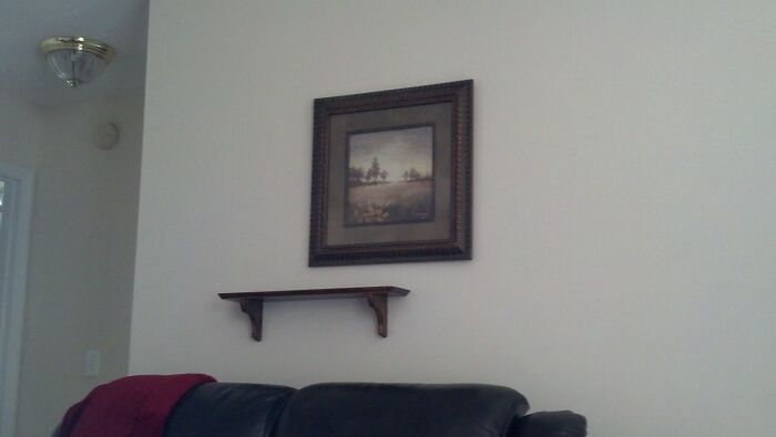 Empty shelf under a framed painting on a white wall, with a sofa and red pillow below, illustrating interior decor choices.