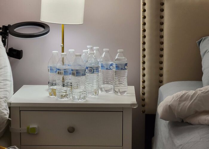 Nightstand cluttered with multiple water bottles, lamp, and bed, related to infuriating wives and girlfriends topic.