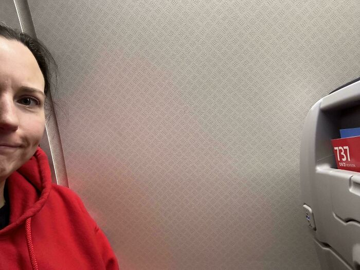 Person in a red hoodie with an irritated expression next to a wall on an airplane.