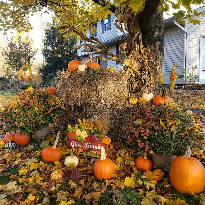 Thanksgiving home decor with pumpkins, hay bales, and a "Give Thanks" sign under a tree, showcasing autumn colors.