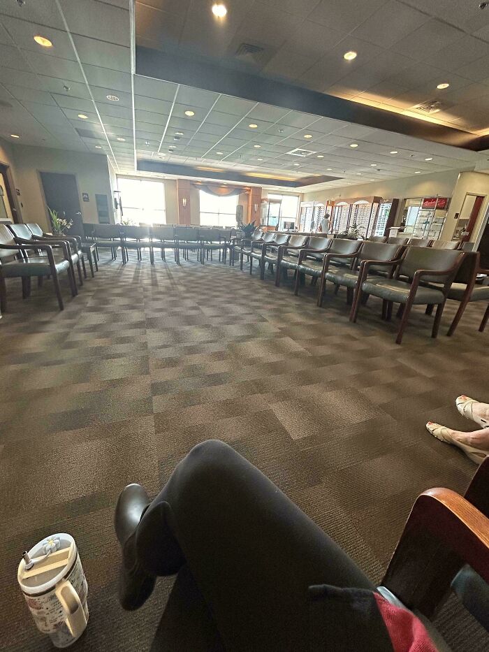 Empty waiting room with chairs in rows, person's legs visible, coffee mug on the floor.