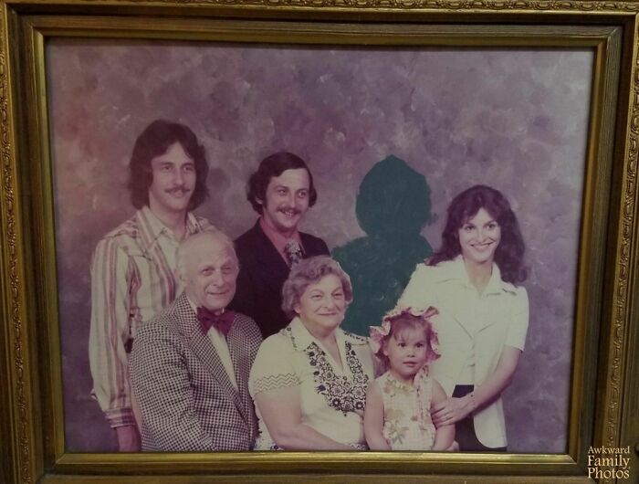 Awkward family photo with a cut-out figure, featuring six people in vintage attire, framed on a wall.