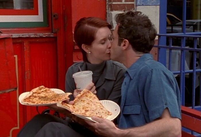 Couple shares a kiss while holding pizza slices, capturing a wholesome ‘90s moment.