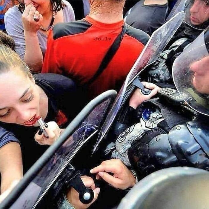 A woman applies lipstick in a crowded protest scene, using a police shield as a mirror. Pics that go hard.